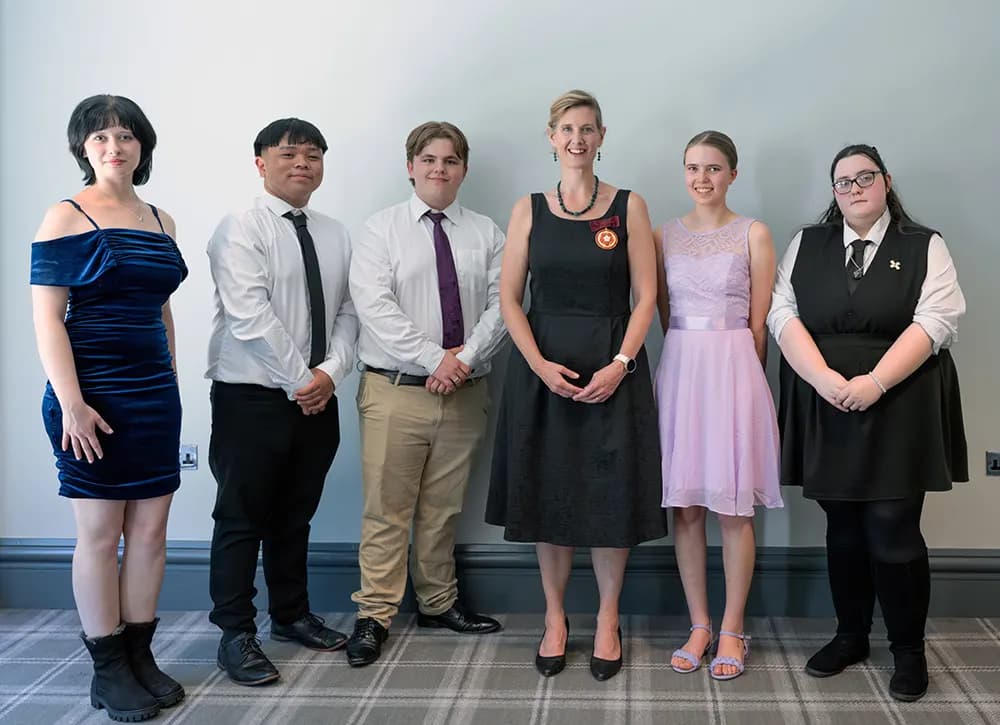 Five young people in formal dress standing in a line with a woman wearing a medal