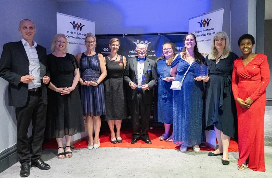 Nine people in evening dress, several holding glass awards, in front of the Pride of Rushmoor Community Awards banners
